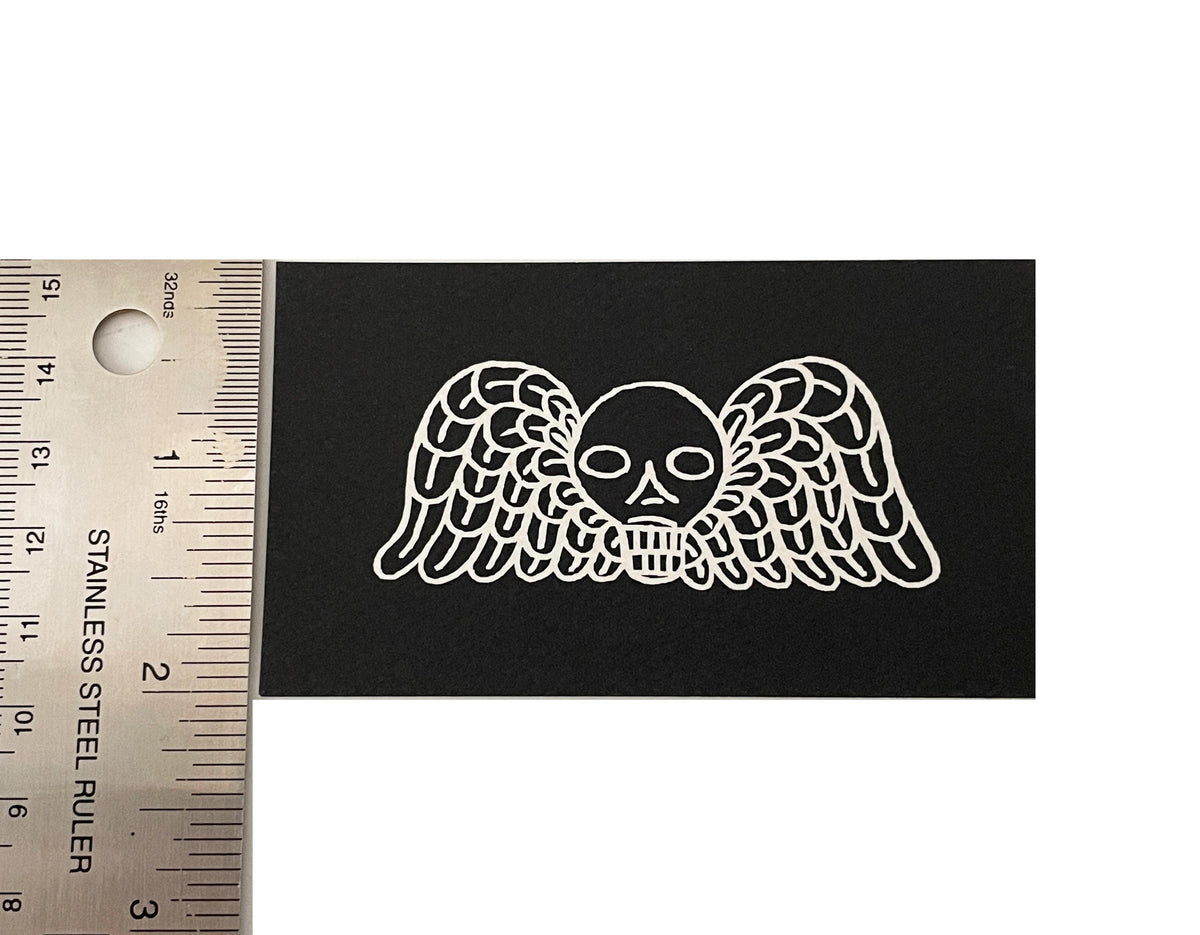 A black rectangular patch featuring a white line-art design of a skull with outstretched wings, shown next to a stainless steel ruler for scale on a white background.